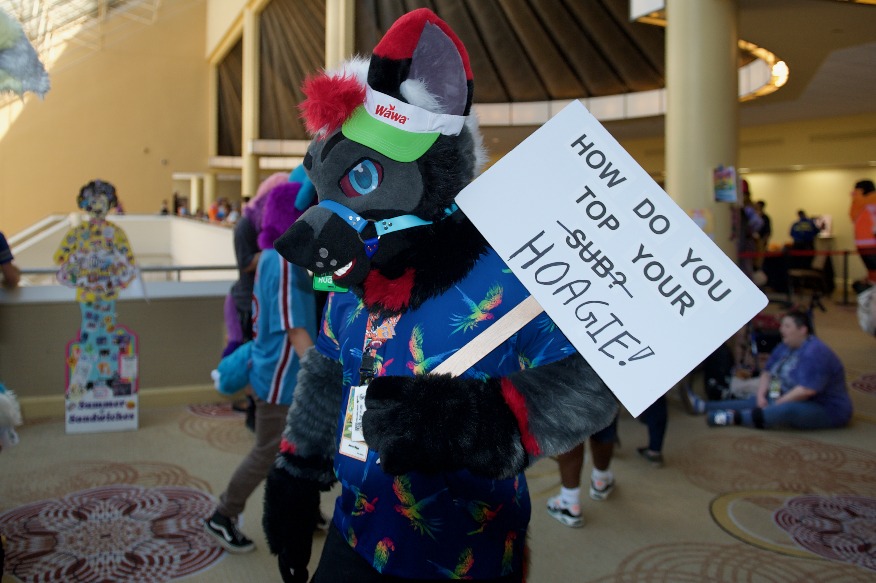 A fursuiter wears a Wawa cap and holds a sign saying 'How do you top your sub?/hoagie!'